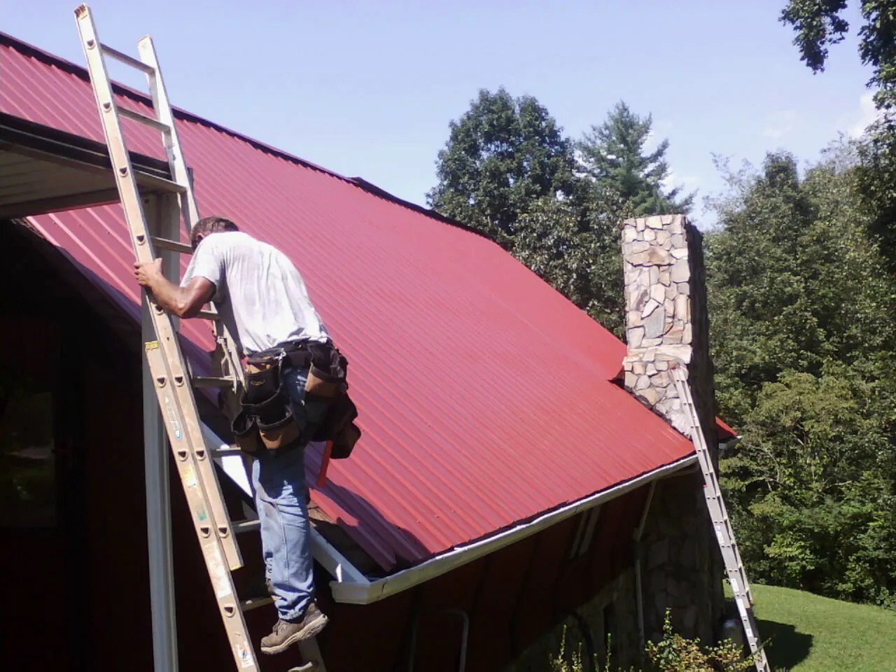 Roofer on a ladder beside a red metal roof and stone chimney — Soffit & Fascia Repair by Roofing Brothers in Fiskeville